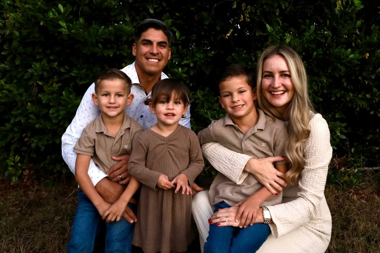Family of five posing together outdoors against a green leafy background, smiling at the camera