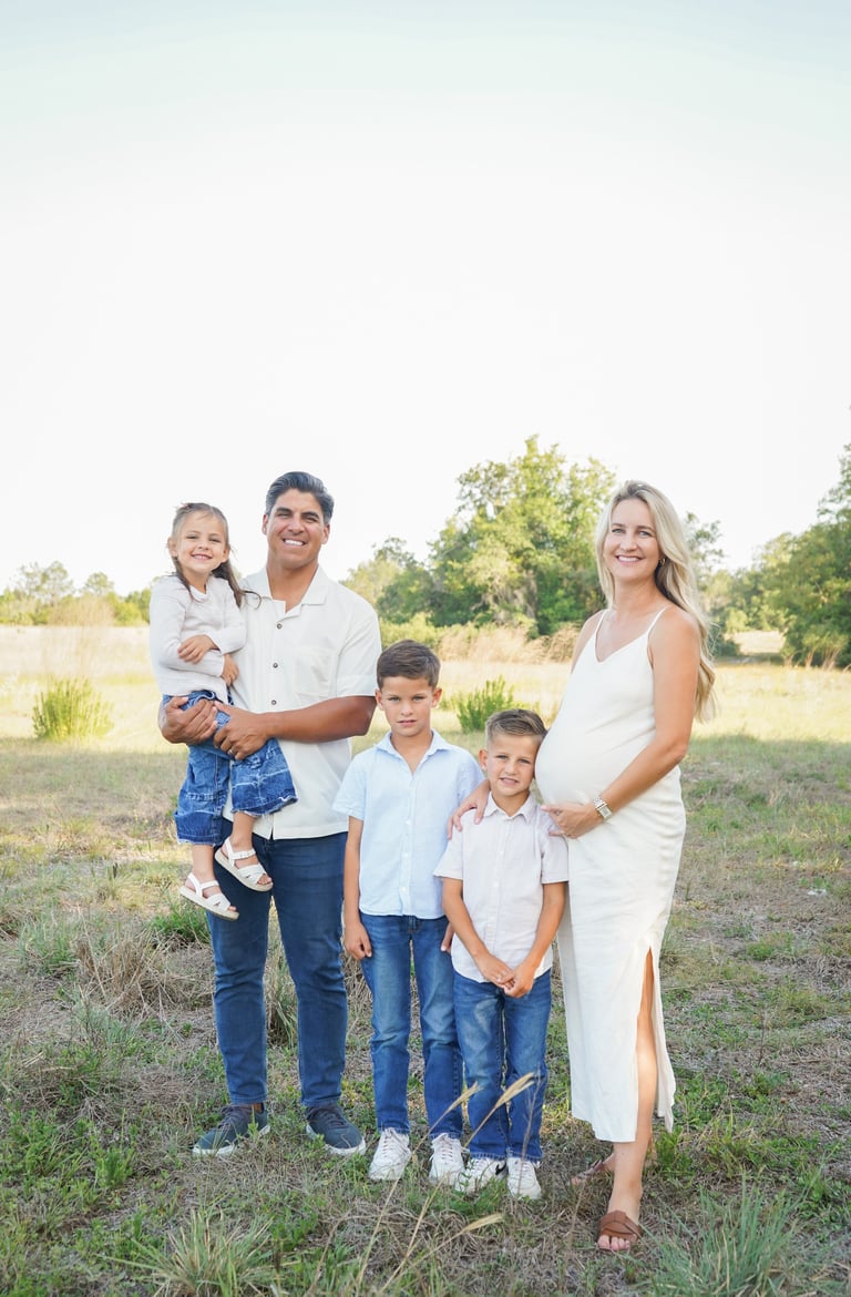 Family of five posing outdoors in a grassy field with trees in background, parents and three children smiling at camera