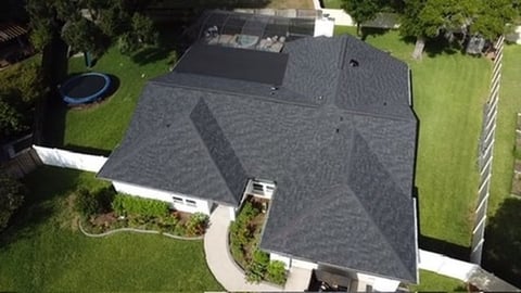 Aerial view of a residential house with gray shingle roof, white trim, and landscaped yard with green lawn and curved driveway