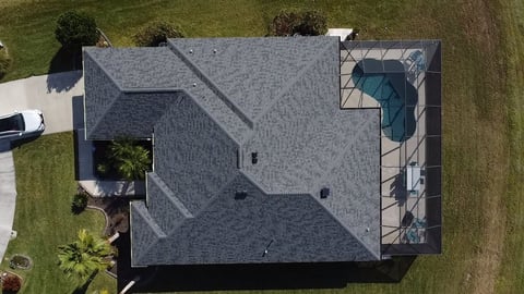 Aerial overhead view of a residential house roof with gray shingles, showing a driveway and pool area surrounded by green lawn and landscaping