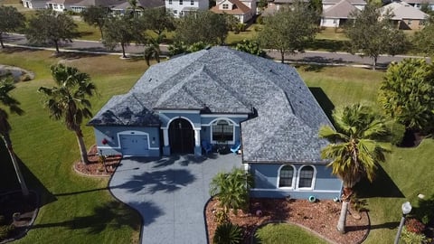 Aerial view of a blue residential home with gray roof, circular driveway, and tropical landscaping on a tree-lined street