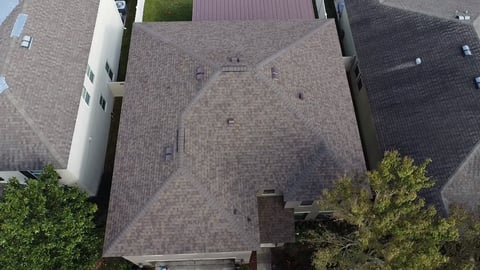 Aerial view of a residential house with gray shingled roof, brick exterior, and surrounding homes visible from above